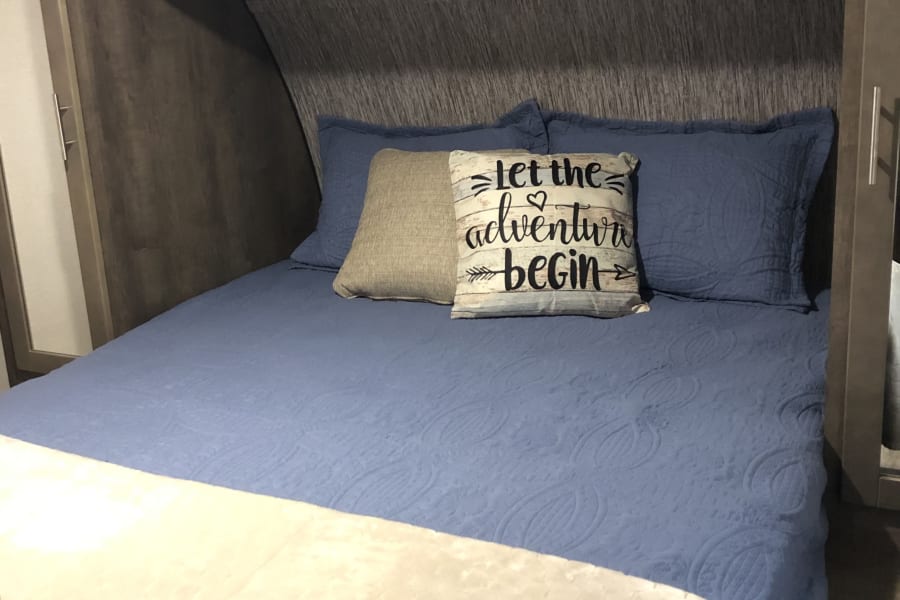 Cozy RV rental bedroom featuring a blue bed with "Let the adventure begin" pillow, and overhead storage.