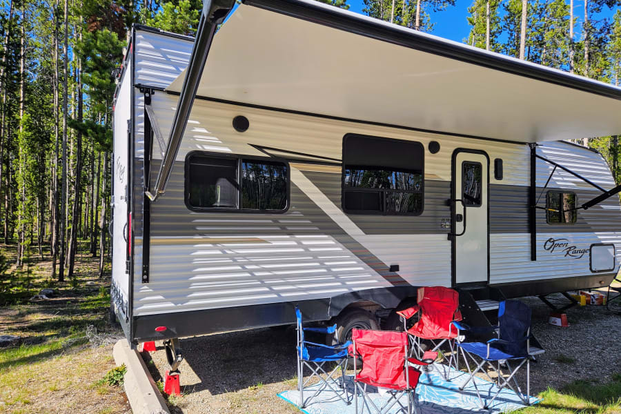 RV with awning, chairs, and mat at a serene forest campsite. Ideal for your next rv rental.