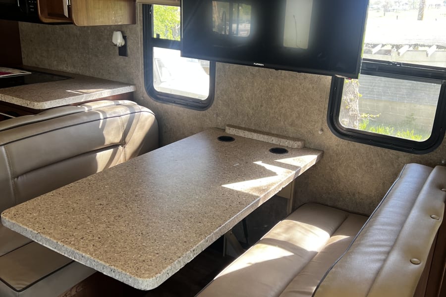 An RV rental interior reveals a dinette with comfy seats, a speckled table, and a wall-mounted TV by sunlit windows.
