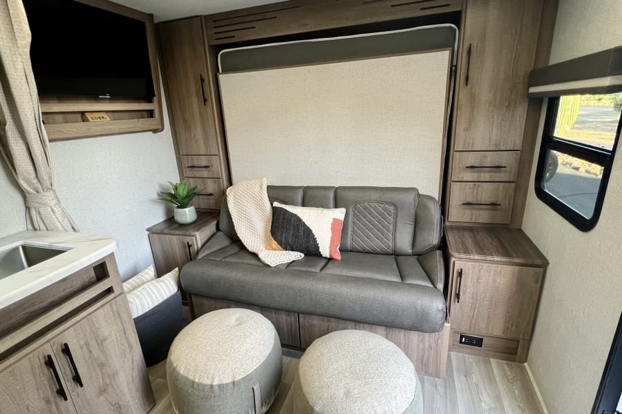 Cozy RV rental interior with a gray sofa, convertible Murphy bed, TV, and built-in cabinetry for comfortable travel.