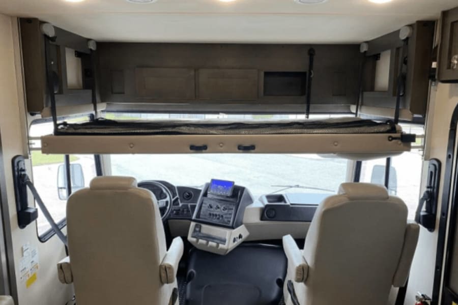 Inside an RV rental: Front cockpit with tan seats, a retractable overhead bunk, and storage. Ready for your motorhome journey.