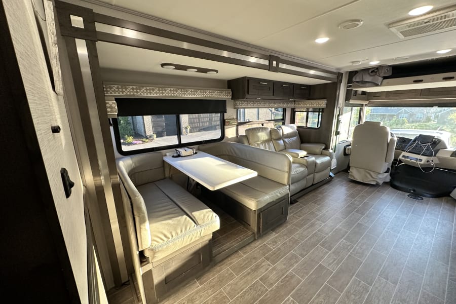 Spacious RV rental interior featuring a dinette, reclining sofa, and cockpit, with modern wood-look tile flooring.
