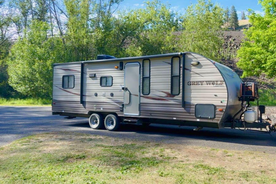 Tan Grey Wolf travel trailer RV rental, parked by green trees & hillside under blue sky. Ready for your outdoor adventure!