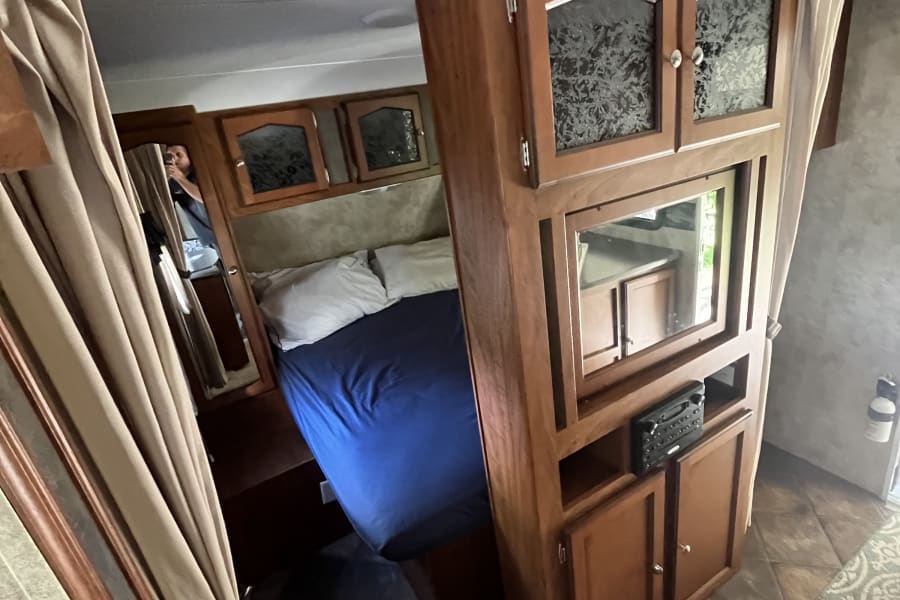 Inside an RV rental: a compact bedroom with a bed, overhead cabinets, and a central storage/entertainment unit.