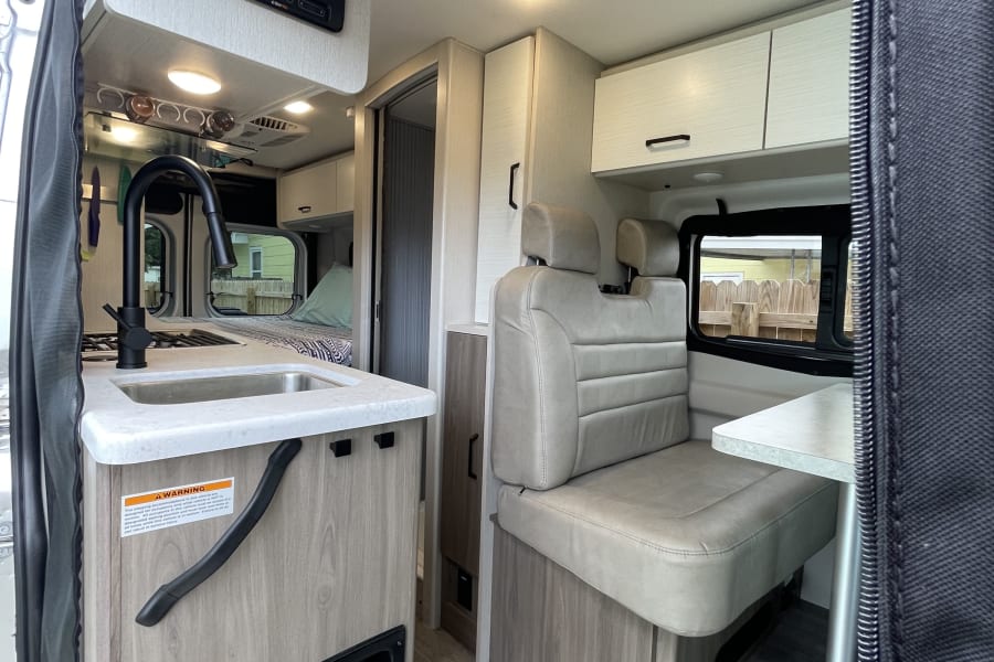 View inside a modern RV rental, featuring a compact kitchen on the left, a light-colored dinette on the right, and a bed area further back.