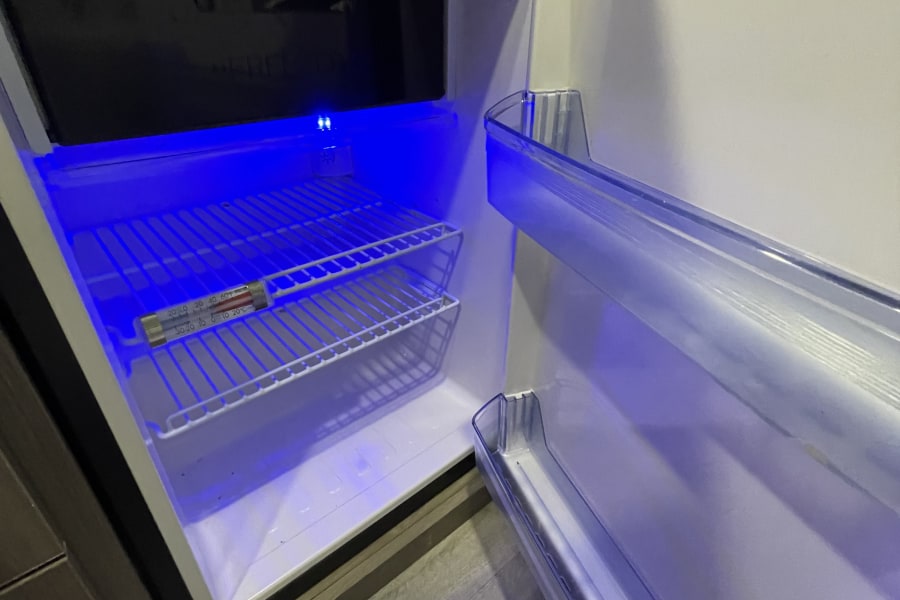 Inside an RV rental refrigerator with a top freezer, illuminated by a blue light. A thermometer rests on a wire shelf.