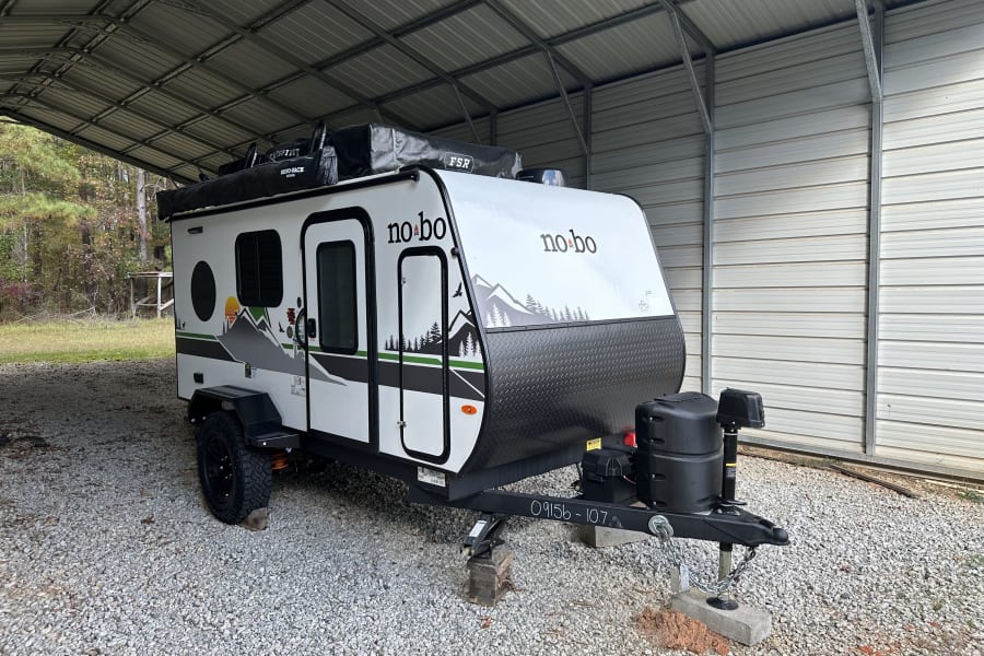This no bo travel trailer, ready for your next outdoor rv rental adventure, is parked on gravel under a metal carport.