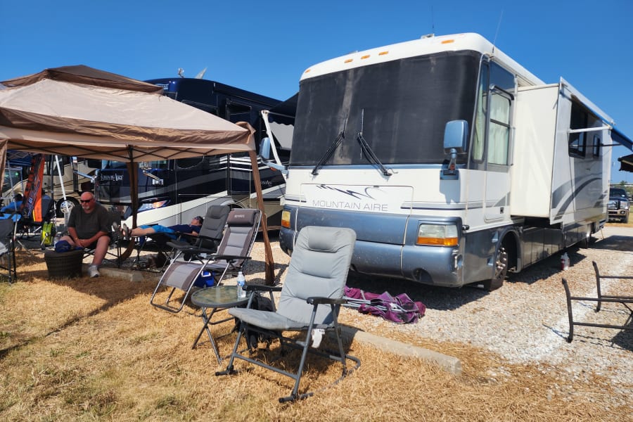 Outdoor relaxation at an RV rental campsite. A Mountain Aire RV is parked, with people relaxing under a canopy.