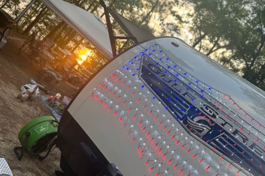 Enjoy a festive RV rental camping experience! This travel trailer with patriotic lights gleams at sunset in a wooded site.