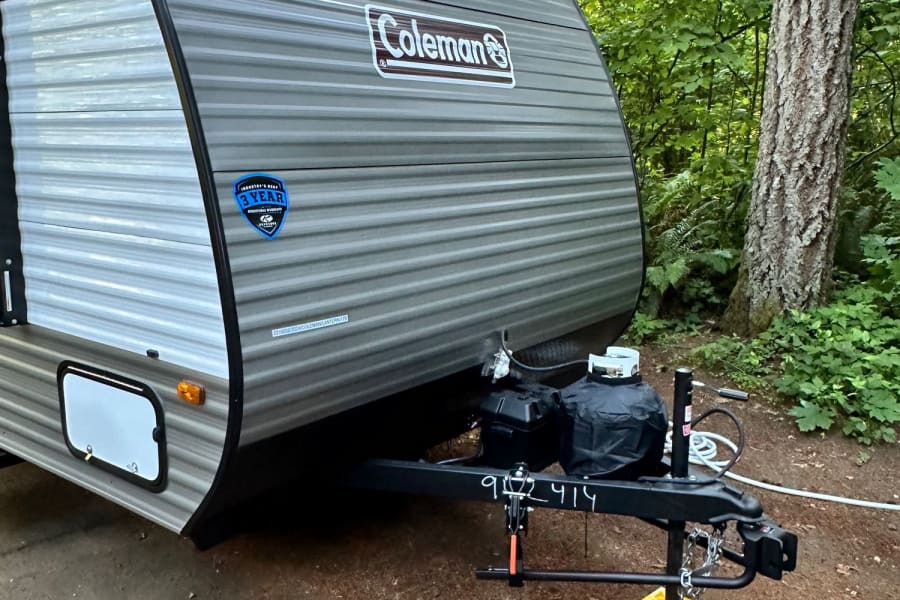 Coleman travel trailer RV rental parked at a serene wooded campsite. Hitch and propane tanks visible at the front.