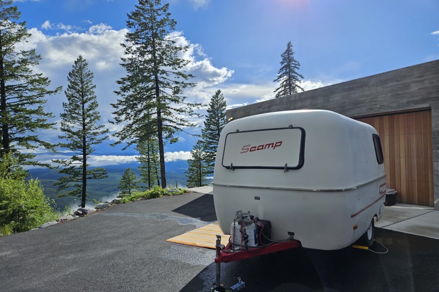 A white Scamp travel trailer, ideal for RV rental, overlooks a misty mountain valley from a modern driveway under a bright sky.