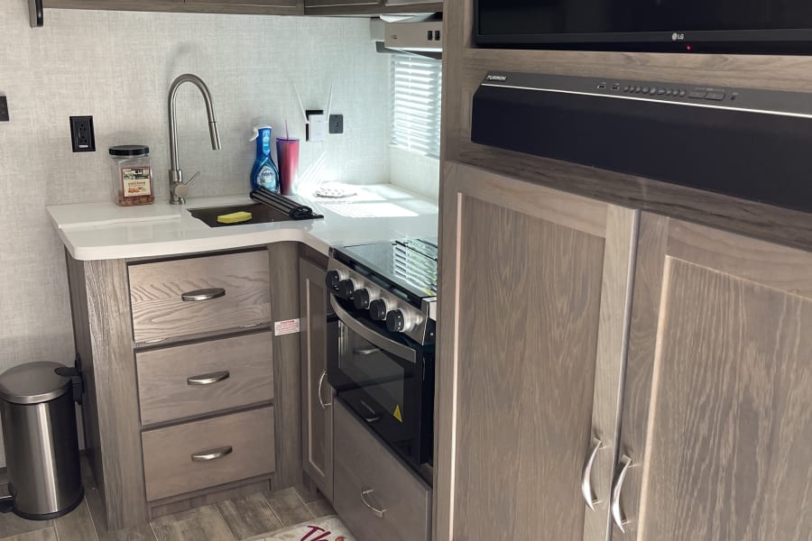 Modern RV rental kitchen featuring grey wood cabinets, sink, oven, and a TV entertainment setup.