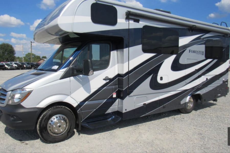 Sleek silver, black & white RV rental motorhome on a gravel lot under blue sky. Note the flat front tire on this Class C camper.