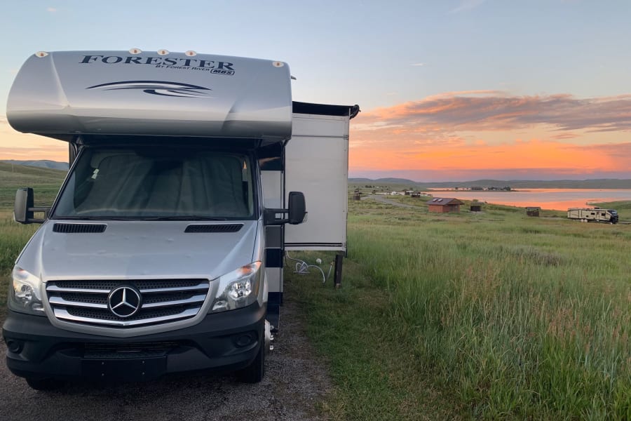 A silver Mercedes-Benz Sprinter RV rental parked by a lake, bathed in the warm glow of a stunning sunset.