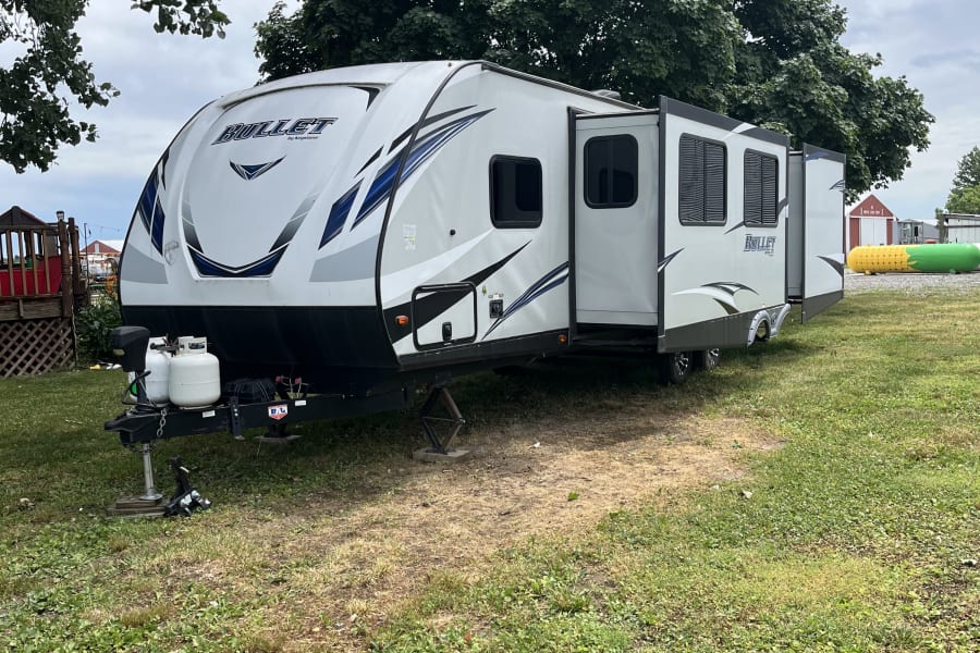 Spacious Bullet by Keystone travel trailer, ideal for your next RV rental getaway. Parked in a grassy area under large trees.