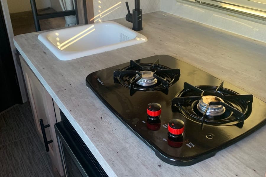 This RV rental kitchenette features a bright sink, black faucet, two-burner stove, and microwave in a compact space.