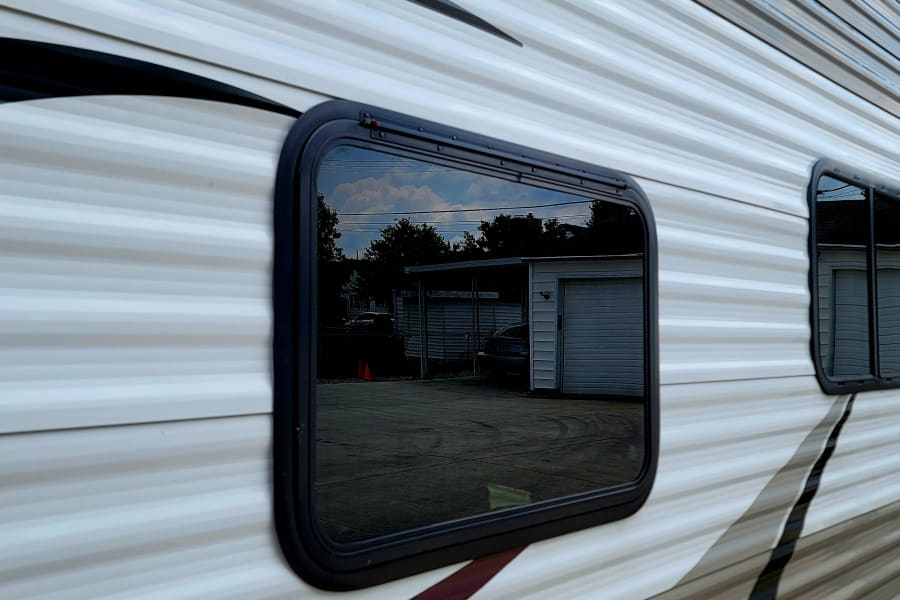 White and gray RV rental exterior, showing horizontal siding and windows reflecting trees, sky, and a garage.