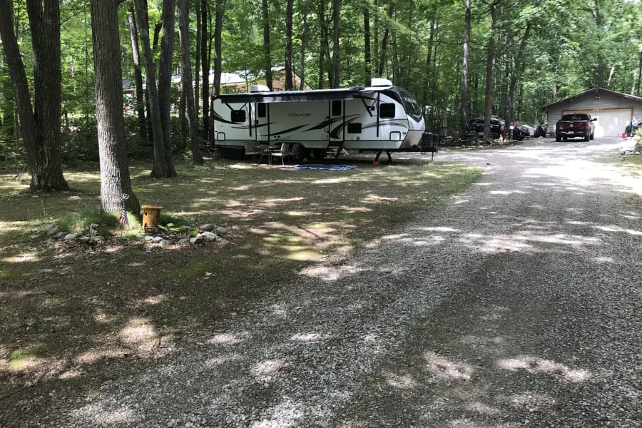 RV rental site: a Cougar travel trailer parked in a serene wooded area with outdoor seating next to a gravel driveway.