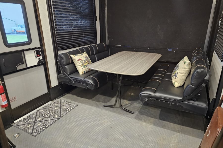 RV rental interior: a cozy dinette with black benches, central table, and decorative pillows. Window, door, and rug visible.
