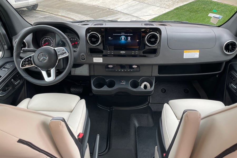 RV rental cockpit view: Mercedes steering wheel, dashboard with infotainment screen, and clean, light-colored front seats.
