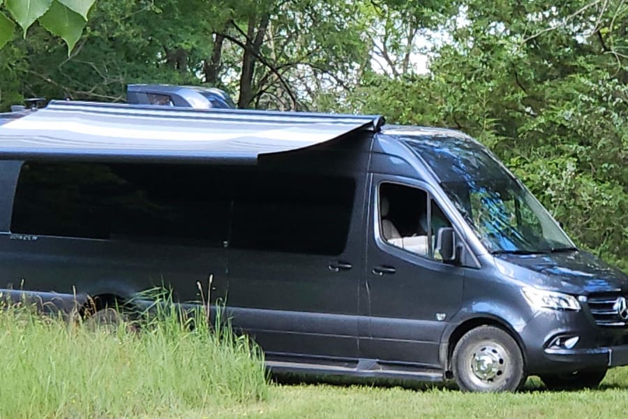Dark gray Mercedes Sprinter camper van RV rental with extended awning, parked in grass outdoors.