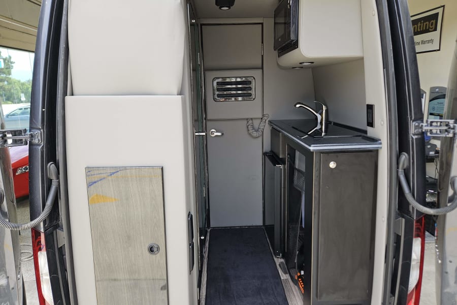 Interior of a modern RV rental. Features a compact kitchen with sink, microwave, and fridge on the right, and a sleeping/seating area on the left.