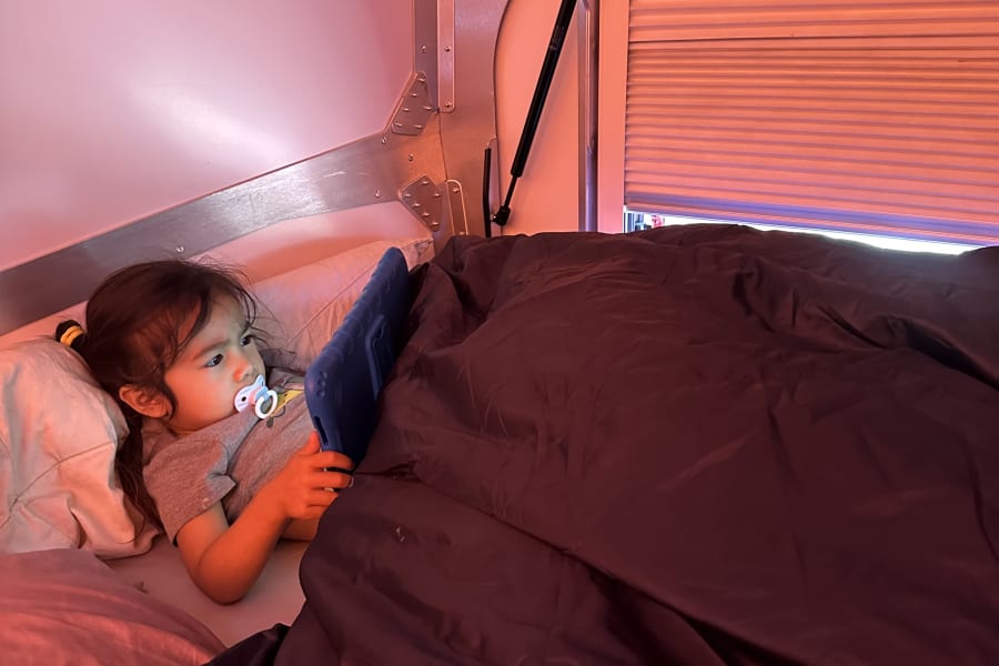 Inside an RV rental, a child in bed uses a tablet under warm light, with an "EXIT/SORTIE" sign nearby.
