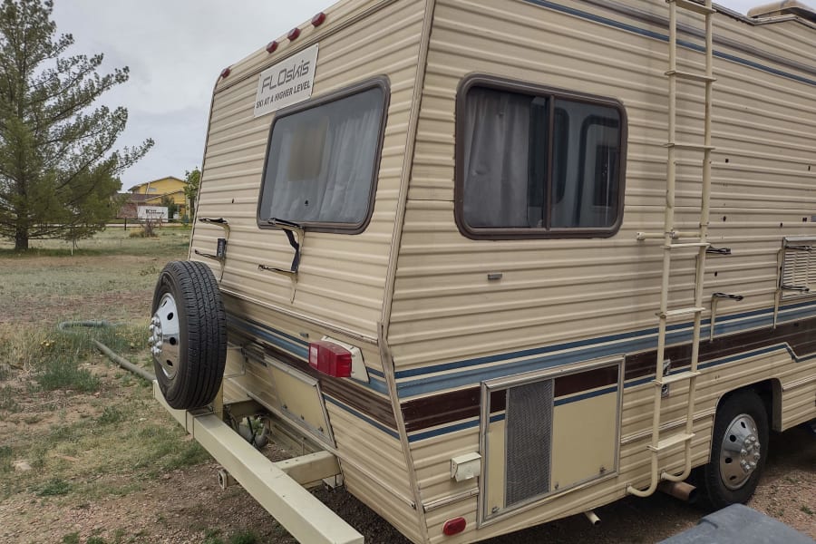 Rear view of a beige RV rental with a spare tire, roof ladder, and "FLOSKIS" sign, parked outdoors.