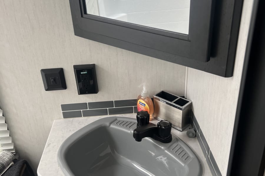 Compact RV rental bathroom featuring a grey sink, black faucet, white counter, and mirror reflecting the shower area.