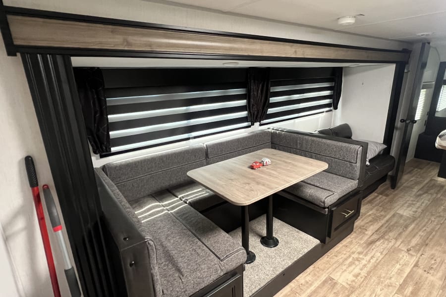 Spacious RV rental dinette featuring grey U-shaped seating, a light wood table, and adjustable window blinds.