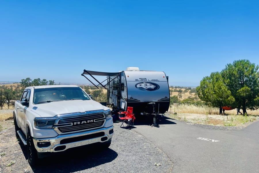 White Ram truck and a travel trailer (RV rental) parked at a serene hilltop campsite under a clear blue sky with rolling hills.