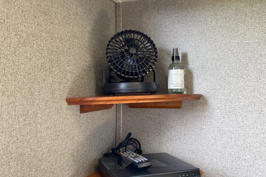 Corner of an RV rental: built-in shelves with a fan, room spray, and DVD player. Speaker overhead for entertainment.