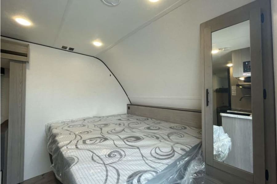 Clean RV rental bedroom featuring a bed with a fresh mattress. Mirrored closet reflects the modern kitchen space.
