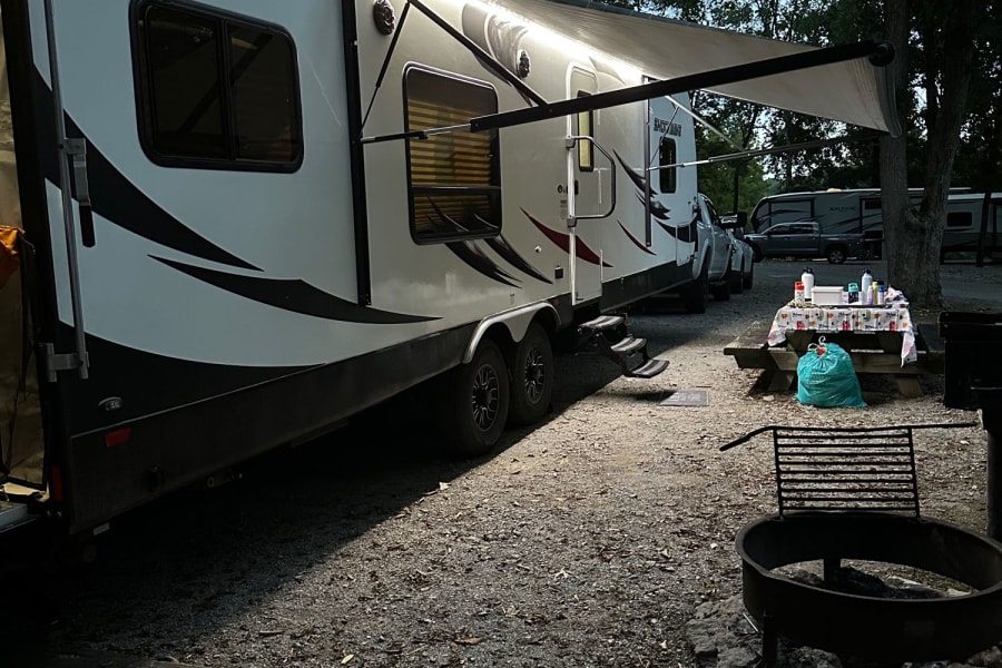 Spacious RV rental at a shaded campsite, featuring an illuminated awning, picnic table, and fire pit for outdoor enjoyment.