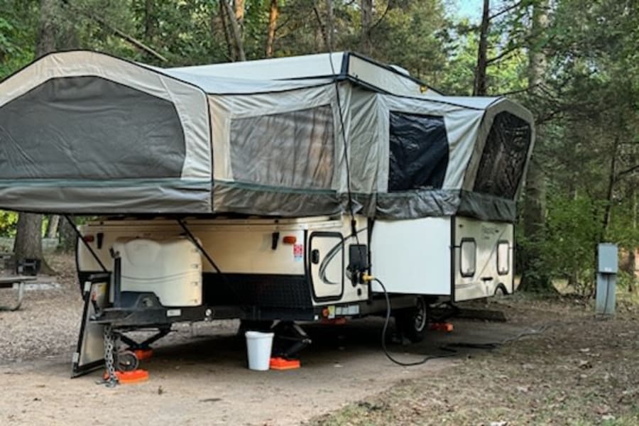 This pop-up RV rental is fully set up at a shady campsite. Its expanded design includes screen windows and utility hookups.