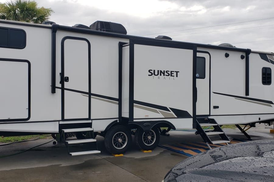 White "Sunset Trail" RV rental travel trailer set up at a site with slides extended, steps down, and utility connections.
