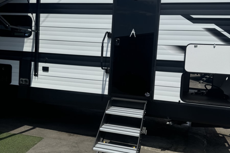 Exterior view of a modern RV rental, featuring tri-fold steps, open storage bays, and a side awning.