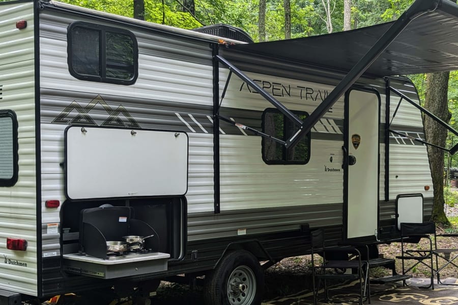 Aspen Trail RV rental ready for camping! Features outdoor kitchen & awning in a lush forest setting.