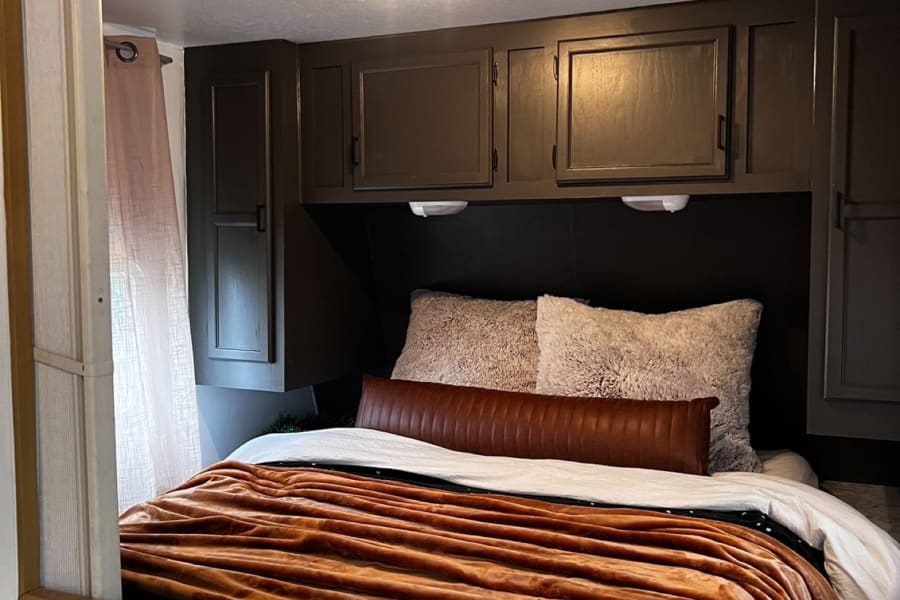 Cozy RV rental bedroom with a comfy bed, plush rust blanket, dark overhead cabinets, and warm reading lights.