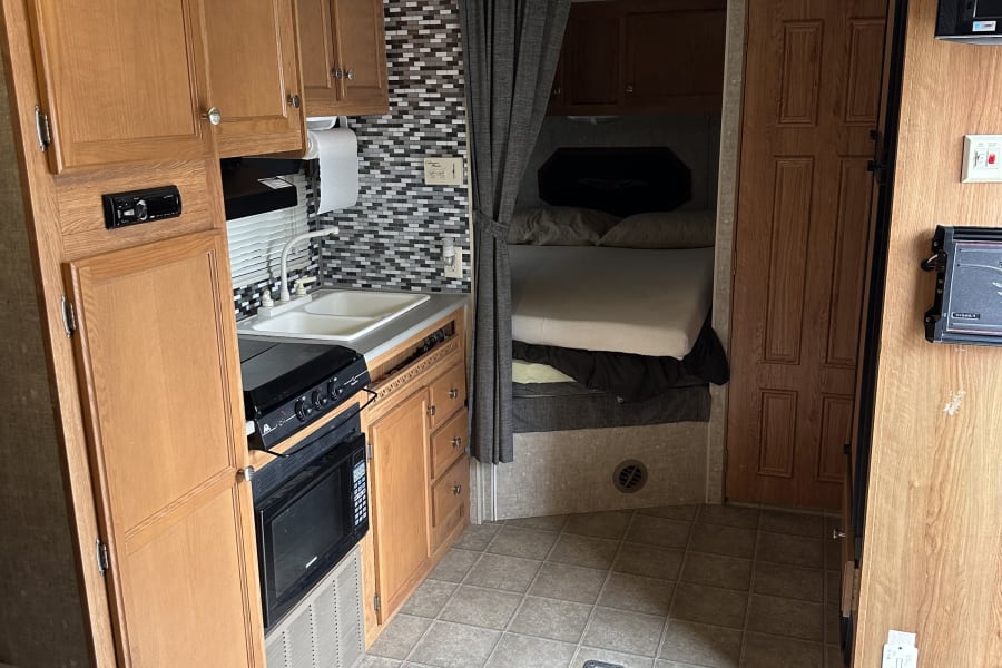 RV rental interior: A compact kitchen with wooden cabinets, sink, and appliances, leading to a private bunk bed area.
