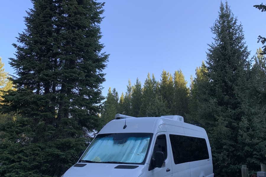 This white Mercedes Sprinter RV rental is parked at a forested campsite, hooked to power for a cozy stay.