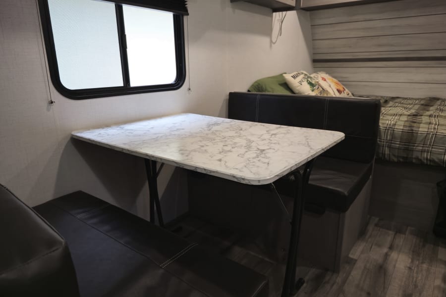 Comfortable RV rental interior featuring a dinette with a white table and black benches, plus a window and bed area.