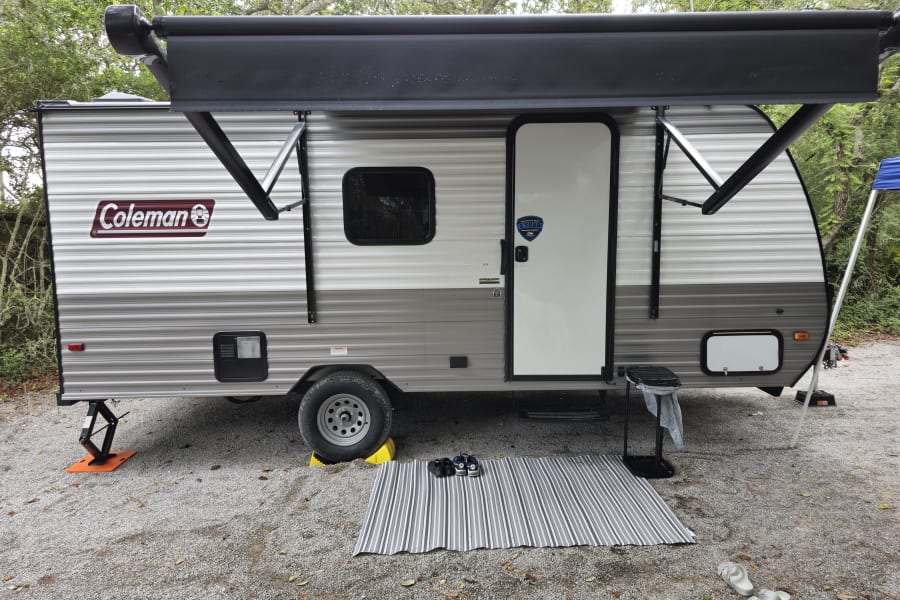 Coleman travel trailer RV rental set up at a gravel campsite with extended awning, ground mat, and outdoor essentials.
