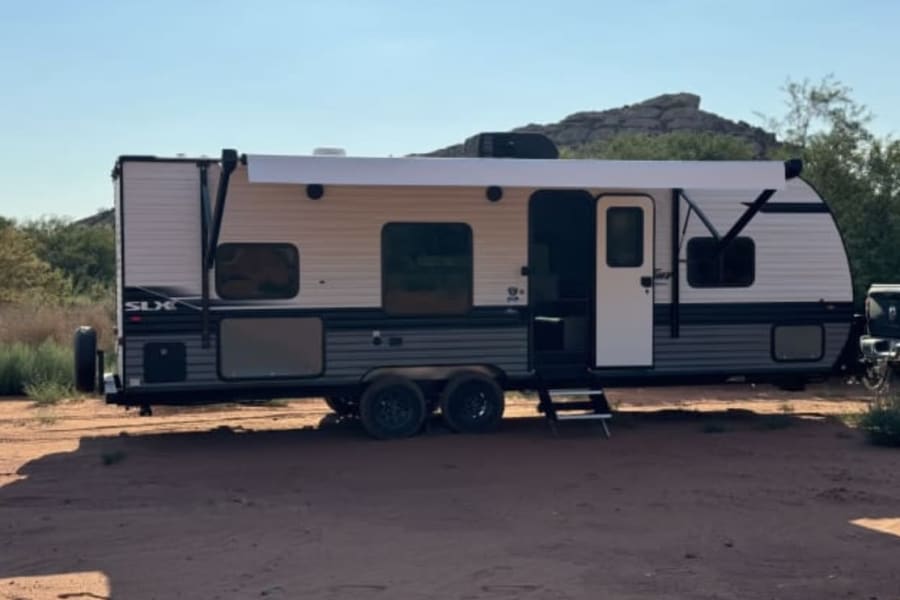 This travel trailer, an SLX model RV rental with awning extended & steps down, is parked on sand with mountains in background.
