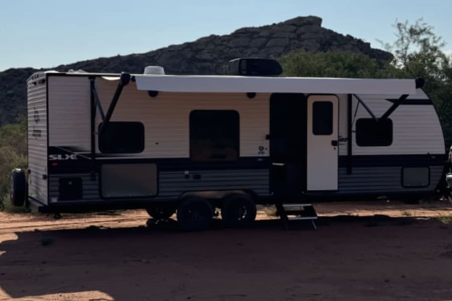 An RV rental is perfectly set up for desert camping, with its awning extended on reddish earth, mountains in the distance.