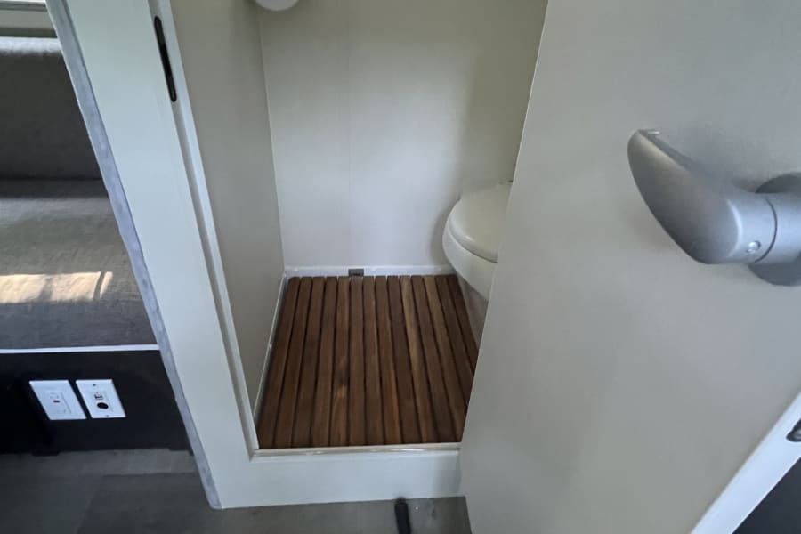 RV rental wet bath, showing compact toilet, wooden shower floor, and circular window with pleated shade.