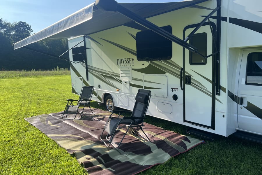 RV rental set up for outdoor living with awning, chairs, and mat on green grass under a clear sky.