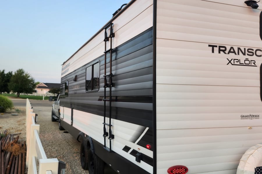 White and black Transcend XPLOR travel trailer, great for RV rental, parked on a residential street with ladder, windows, and a truck.