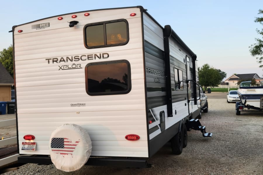 A Grand Design Transcend XPL&Ocirc;R travel trailer, perfect for an RV rental, is parked on a gravel driveway next to a boat.