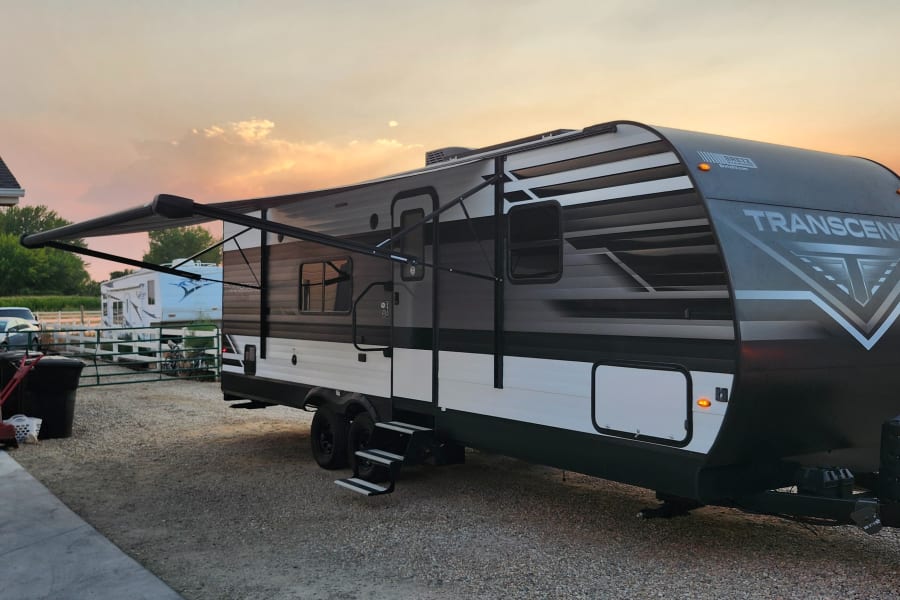 A modern Transcend travel trailer, great for an rv rental, with its awning extended on a gravel lot at sunset.